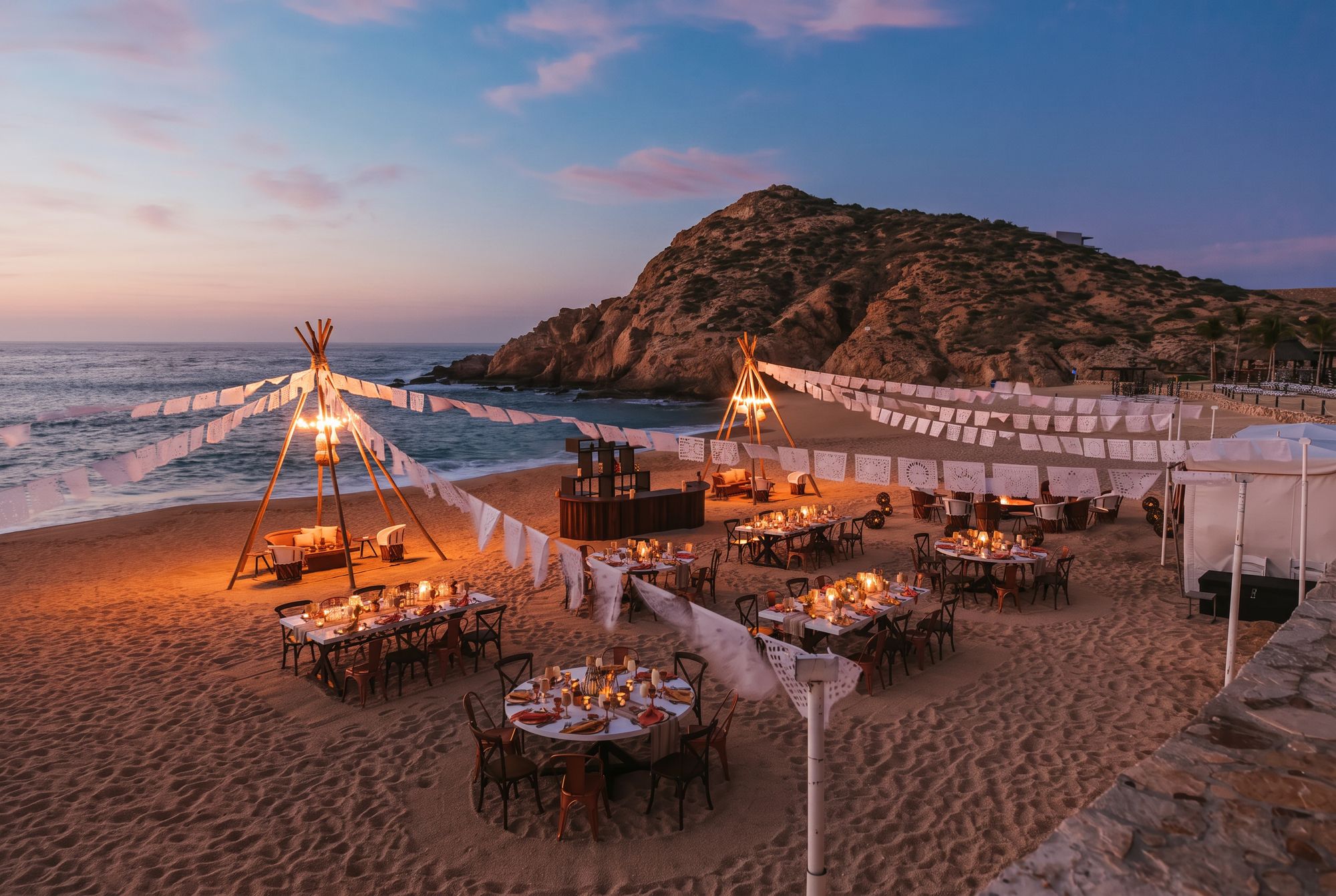Candlelit beach dinner setup at sunset with papel picado banners and teepee lanterns along the Sea of Cortez